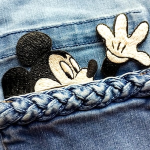 Può includere: Primo piano di una tasca di jeans blu chiaro con un bordo intrecciato. Un disegno ricamato di Topolino emerge dalla tasca, con la sua testa e una mano visibili. Il disegno presenta fili neri, bianchi e beige.