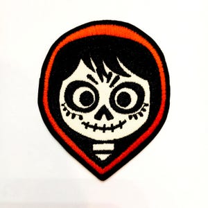 Puede incluir: Parche bordado con un diseño de calavera estilizada con grandes ojos negros, una boca sonriente y cabello negro. El parche tiene un borde rojo y está sobre un fondo blanco. El diseño recuerda a un personaje de la película Coco.
