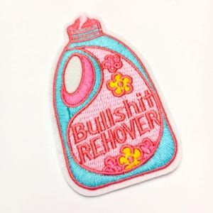 May include: Embroidered patch in the shape of a detergent bottle, featuring the text "Bullshit Remover" in pink. The patch has a pink and teal color scheme with floral accents.