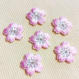Könnte beinhalten: Sechs gestickte Aufnäher in Form von rosa Blüten mit weißen und silbernen Zentren. Die Blütenblätter haben einen Farbverlauf von Rosa, mit einem helleren Farbton an den Rändern. Die Aufnäher liegen auf einem hellbeigen Stoff.