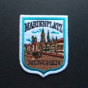 Op de afbeelding: Een witte geborduurde patch met een blauwe rand. De patch toont een stadsbeeld met gebouwen en een kerktoren. De tekst "MARIENPLATZ MÜNCHEN" is in zwart geborduurd.