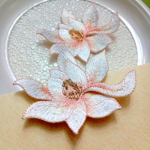 May include: Two embroidered flower patches featuring white petals with soft pink shading and gold-coloured centres. The floral designs have detailed stitching throughout the petals, creating a textured appearance for these decorative fabric appliqués.