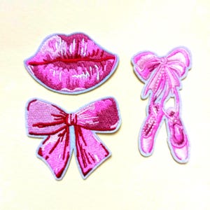 Op de afbeelding: Drie geborduurde patches: een paar roze lippen, een roze strik en een paar roze balletschoenen. Elke patch heeft een lichtblauwe rand. De lippen en de strik zijn in een donkerder roze, terwijl de balletschoenen lichter zijn.