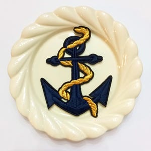 May include: Navy blue anchor patch with gold rope detail, displayed on a cream-colored, scalloped-edge plate. The nautical-themed patch is embroidered with a detailed design. The plate has a glossy finish and a decorative, wavy rim.
