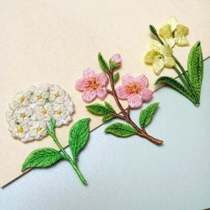 May include: Embroidered floral patches featuring a white hydrangea, pink cherry blossoms, and yellow daffodils. Each flower has green leaves and stems, with detailed stitching. The patches are arranged on a light beige and light blue background.