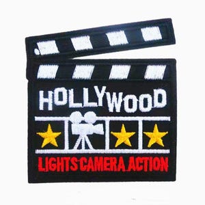 May include: A black and white embroidered patch in the shape of a film clapperboard. The patch features the word "HOLLYWOOD" in white, a film camera graphic, three yellow stars, and the text "LIGHTS CAMERA ACTION" in red.