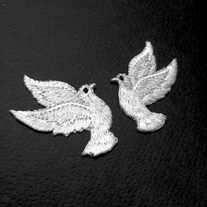 Puede incluir: Dos parches bordados de palomas blancas con costuras detalladas y pequeños ojos negros. Cada ave está representada en posición de vuelo con las alas extendidas, mostrando la textura del hilo sobre un fondo oscuro.