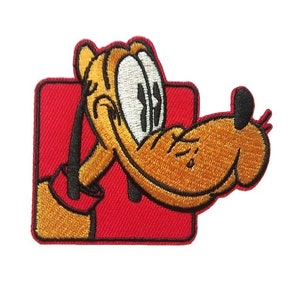 Op de afbeelding: Een geborduurde strijk-op patch met een cartoon afbeelding van Pluto, een bruine en gele hond met grote ogen, die uit een rood vierkantje kijkt.