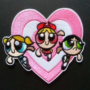 May include: An embroidered patch featuring three cartoon characters with large eyes, positioned in front of a layered pink and white heart shape. The characters have blonde, orange, and black hair, wearing blue, red, and green outfits respectively.