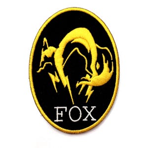 Pode incluir: Patch bordado com a silhueta de uma raposa amarela sobre um fundo oval preto. A palavra "FOX" é bordada em letras brancas. O patch tem uma borda amarela.