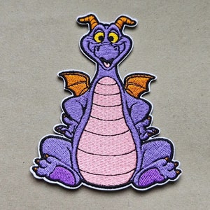 Könnte beinhalten: Gestickter Aufnäher mit einem lila Drachen-Cartoon-Charakter mit orangefarbenen Flügeln und einem rosa Bauch.