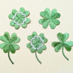 May include: Five green embroidered shamrock patches. Two patches have white and green detailing in the center. The shamrocks have varying numbers of leaves and stems. The patches are on a light beige background.