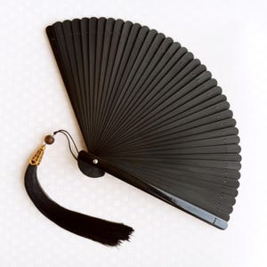 Op de afbeelding: Een zwarte opvouwbare handventilator met een kwast. De ventilator is gemaakt van donker hout en heeft een decoratieve kwast met een goudkleurig filigraan accent. De ventilator is open en toont zijn vele verticale lamellen.
