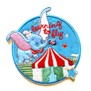 May include: Embroidered patch depicting Dumbo the elephant flying, with a circus tent and the words "Learning to Fly." The patch is circular, with a light blue background and a star-shaped accent.