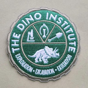 May include: A green and white embroidered patch with the text "The Dino Institute" in a circle around a white dinosaur silhouette. The text "Exploration - Excavation - Exultation" is written below the dinosaur.