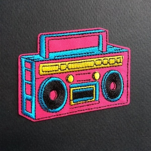May include: A pink, blue, and yellow embroidered patch of a boombox cassette player. The boombox has a yellow cassette player, two black speakers, and a blue handle.