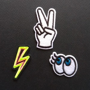 May include: Four iron-on patches with a white background and black outlines. The patches include a peace sign, a lightning bolt, and two cartoon eyes.