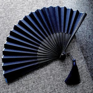 May include: A dark blue folding hand fan with a pleated fabric surface and a matching dark blue tassel attached to the base. The fan features a series of dark, rigid ribs that radiate from the central pivot point, creating a structured, semi-circular shape.