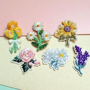 Puede incluir: Un conjunto de parches bordados con flores, incluyendo girasoles, margaritas, una rosa y lavanda. Los parches presentan costuras detalladas en varios colores, como amarillo, blanco, rosa y morado, con hojas y tallos verdes. Ideales para decorar ropa o accesorios.