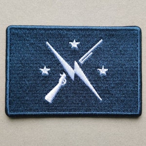 May include: A dark blue embroidered patch with a white lightning bolt design, two crossed rifles, and four stars.