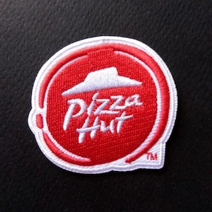 May include: A red and white embroidered patch featuring the Pizza Hut logo. The logo shows a white pizza box with a red roof and the words "Pizza Hut" in white.
