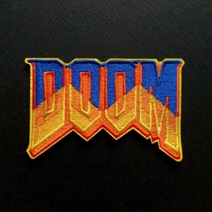 May include: Embroidered patch with the word "DOOM" in a striking, stylized font. The patch uses blue, orange, and yellow thread. The design is outlined with yellow stitching, creating a bold visual effect. The patch is likely for clothing or accessories, and is suitable for sewing.