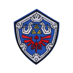 May include: An embroidered patch featuring the iconic Triforce symbol from the Legend of Zelda video game series. The Triforce is surrounded by a stylised shield design, in blue and white.