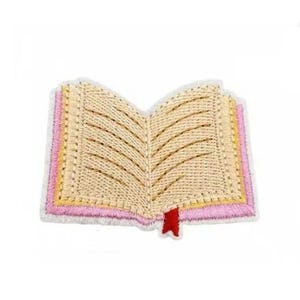 Op de afbeelding: Een beige geborduurde boek patch met een roze en gele rand en een rood bladwijzer. Het boek is open en de pagina's zijn zichtbaar.