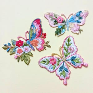 Op de afbeelding: Drie geborduurde vlinderpatches met bloemmotieven. De vlinders hebben een verscheidenheid aan kleuren, waaronder roze, blauw, groen en oranje. Elke vlinder heeft een uniek patroon en is versierd met bloemen en bladeren.