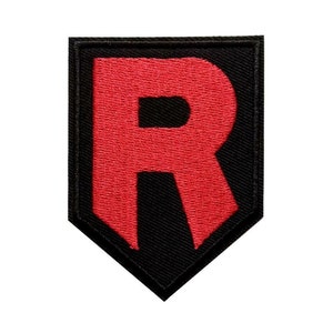 May include: A black and red embroidered patch featuring a large red letter "R" on a black background. The patch has a shield-like shape with a pointed bottom and a black border.