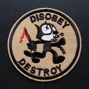 May include: A beige round patch with black embroidery. The patch features a cartoon cat holding a red knife and a chain. The text "DISOBEY DESTROY" is embroidered in black around the cat.