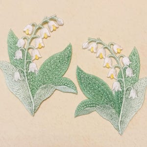 Könnte beinhalten: Zwei gestickte Maiglöckchen-Patches. Jeder Patch zeigt weiße, glockenförmige Blüten mit gelben Zentren und grünen Blättern. Die Patches sind mit verschiedenen Grüntönen und weißem Faden detailliert, wodurch ein strukturiertes Aussehen entsteht.