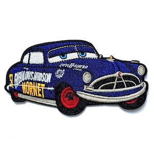 Puede incluir: Un parche de tela bordado con forma de coche antiguo azul con ojos blancos en el parabrisas. El lateral presenta el texto Fabulous Hudson Hornet en letras amarillas y blancas. El coche tiene ruedas rojas y un parachoques delantero plateado.
