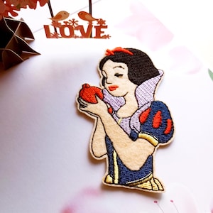 May include: Embroidered patch featuring Snow White holding a red apple. The patch has a cream-colored background with a blue dress, red accents, and a black bob. The patch is a whimsical depiction of the classic fairytale character.
