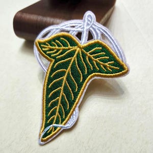 May include: An embroidered patch shaped like a three-lobed leaf with deep green fabric, gold thread veins, and a white stitched border. The design features a stylised stem loop at the top, creating a distinct botanical shape with detailed thread work.