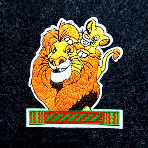 May include: An embroidered patch featuring a large orange lion with a full mane and a smaller yellow lion cub perched on its head. Below the lions is a rectangular bar with a red and green diagonal striped pattern and a white border.