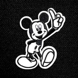 Pode incluir: Patch bordado com Mickey Mouse em preto, branco e bege. Mickey está sorrindo e apontando para cima com o dedo indicador. O patch tem uma borda branca sobre um fundo preto texturizado.