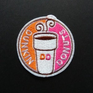Op de afbeelding: Een ronde, witte, geborduurde patch met een roze en oranje achtergrond. De patch heeft een koffiebeker met stoom die eruit opstijgt en de tekst "Dunkin' Donuts" in een cirkelvormig ontwerp.