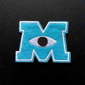 May include: A blue felt patch with a white outline in the shape of the letter "M". A white eye with a black pupil is centred in the "M".