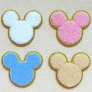May include: Four fuzzy fabric Mickey Mouse head appliques with gold trim. The appliques are white, pink, light blue, and beige.