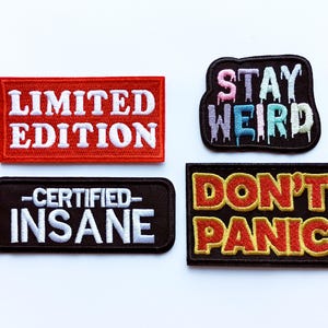 May include: Four embroidered patches with text. The top left patch is red with white text that reads "LIMITED EDITION." The top right patch is black with colorful text that reads "STAY WEIRD." The bottom left patch is black with white text that reads "CERTIFIED INSANE." The bottom right patch is black with yellow and red text that reads "DON'T PANIC."