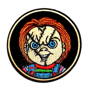 May include: A circular embroidered patch featuring a stylised character with bright orange hair, pale skin, and prominent facial scars. The character has piercing blue eyes and a wide grin, wearing a blue overall-style outfit with a green shirt.