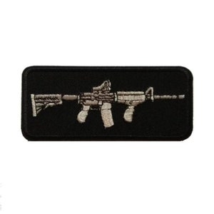 Puede incluir: Un parche bordado negro con un contorno blanco de un rifle.