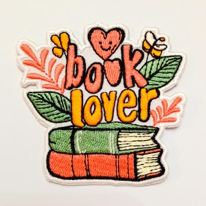 Puede incluir: Parche bordado con las palabras "book lover" en naranja y rosa, con un corazón sonriente y detalles florales. Dos libros apilados en la parte inferior, uno verde y otro naranja.