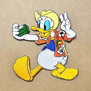 May include: An embroidered patch featuring a cartoon duck wearing a yellow hat and an orange floral shirt. The duck has white feathers, a yellow beak, and yellow feet, and is holding a small green rectangular object while making a peace sign.
