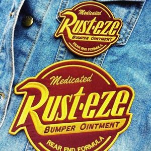 Pode incluir: Dois patches bordados com o texto "Medicated Rust-eze Bumper Ointment Rear End Formula" em amarelo e vermelho. Os patches estão em uma jaqueta jeans azul.