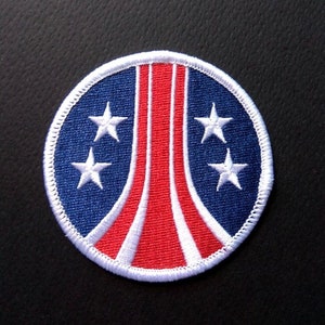 Może przedstawiać: Naszywka haftowana w kolorze białym, czerwonym i niebieskim z geometrycznym wzorem rakiety i trzema gwiazdami. Naszywka jest okrągła i ma białą obwódkę.