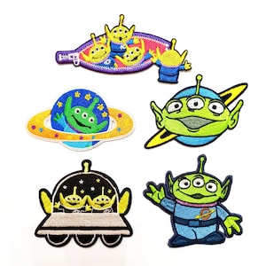 Op de afbeelding: Een verzameling geborduurde patches met de groene, drieoogige aliens uit de Toy Story-franchise. Ontwerpen omvatten aliens in een ruimteschip, op een planeet en in verschillende poses. De patches hebben gele, blauwe en groene accenten.
