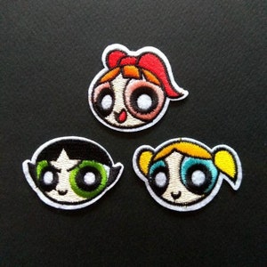 May include: Three embroidered iron-on patches featuring the faces of the Powerpuff Girls: Blossom, Bubbles, and Buttercup. The patches are white with colourful details.