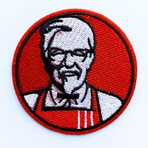 Op de afbeelding: Geborduurde ronde patch met een portret van een man met wit haar, een bril en een sik, tegen een rode achtergrond. De man draagt een vlinderdas en een rode schort met witte strepen. De patch heeft een zwarte rand.